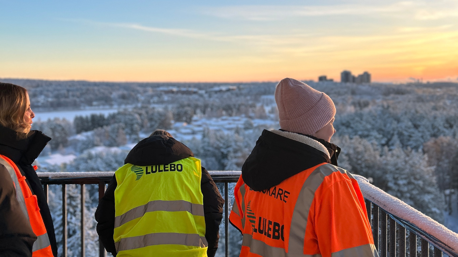 Tre personer tittar ut från en balkong högst upp i ena lägenhetshuset på Lingonstigen 237. I bakgru